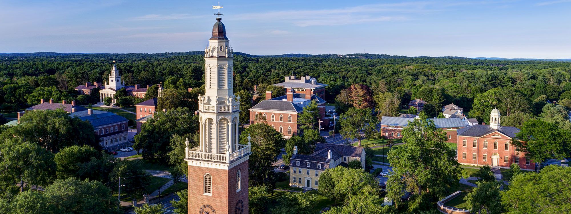 An aerial view of a historic campus with a prominent clock tower, surrounded by lush greenery and multiple brick buildings.