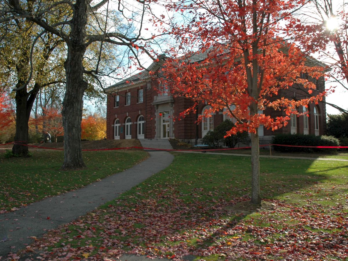 A brick building is surrounded by autumn trees with vibrant red and orange leaves, set beside a walkway and grassy area.