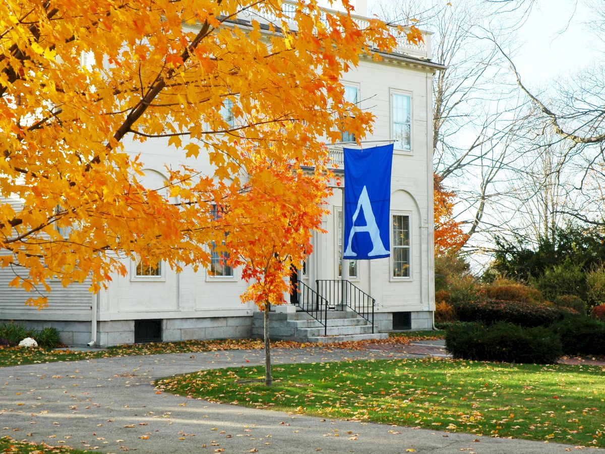 A brick pathway leads to a building with a blue "A" banner, surrounded by autumn trees and a grassy area.