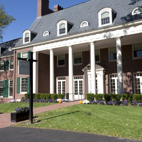 A large brick building with white columns, dormer windows, and a well-maintained lawn is pictured.
