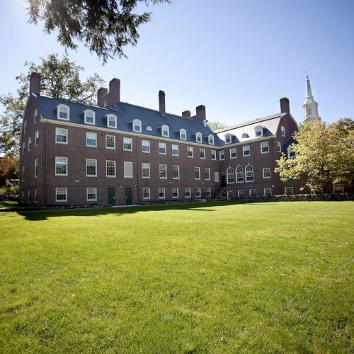 A large brick building with multiple windows is set against a clear blue sky and surrounded by green grass and trees.
