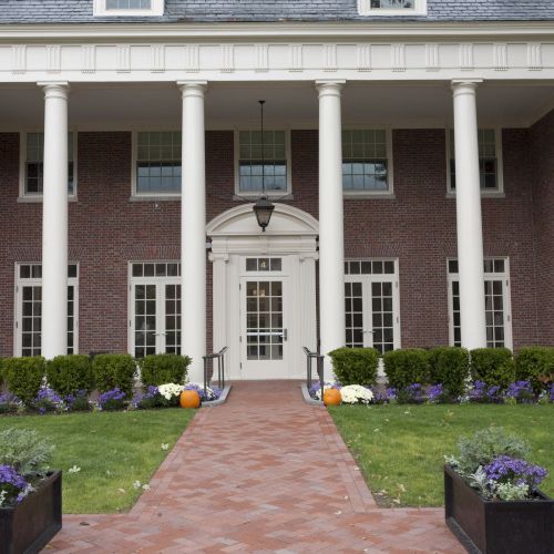 The image shows the entrance of a brick building with large white columns, potted plants, and a pathway, resembling an institutional facade.