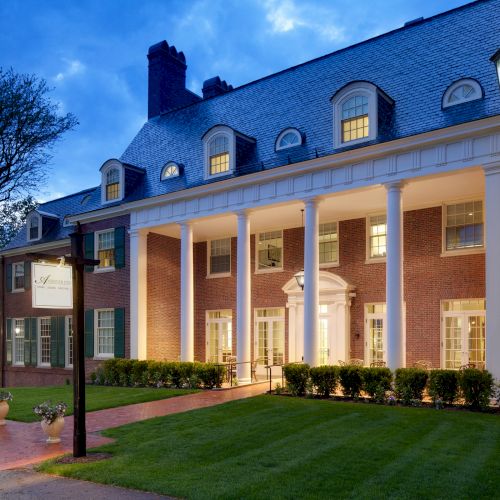 A brick building with large white columns and glowing windows at dusk, surrounded by manicured grass and lit street lamps.