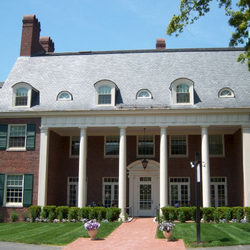 A large brick building with white columns and green shutters, featuring a landscaped front with a walkway and potted plants.