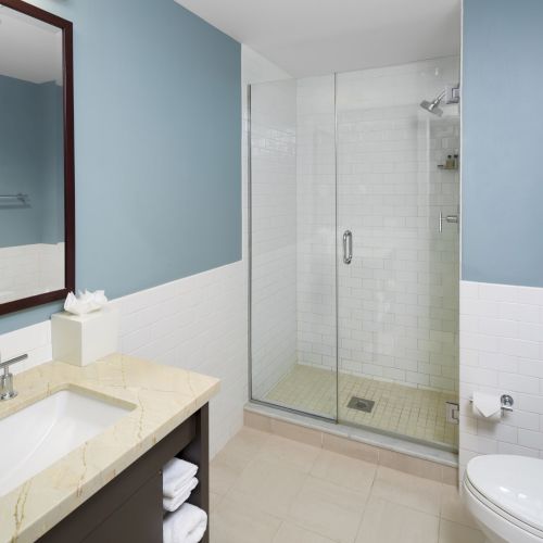 A modern bathroom with a sink, mirror, and glass shower. The walls are light blue, and there's a toilet to the right.