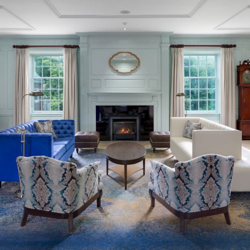 The image shows an elegant living room with a fireplace, blue and white seating, a grandfather clock, and large windows with curtains.