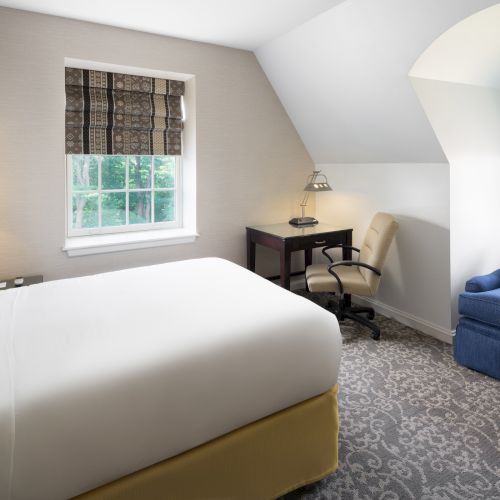 A cozy bedroom with a bed, desk, chair, lamp, and a small sofa near a window; neutral tones and patterned curtains enhance the comfort.