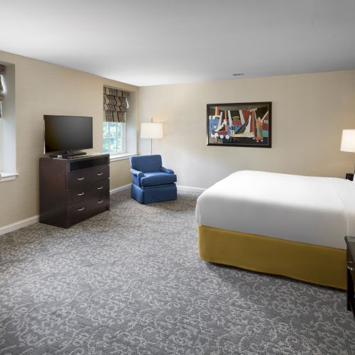 The image shows a hotel room with a bed, TV, desk, chair, armchair, lamps, and a window with patterned curtains.
