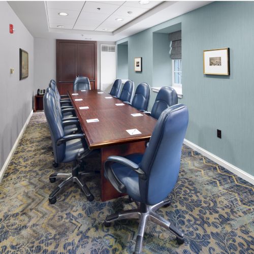 A conference room with a long table, blue chairs, and artwork on light blue walls; the carpet features a blue and gray pattern.