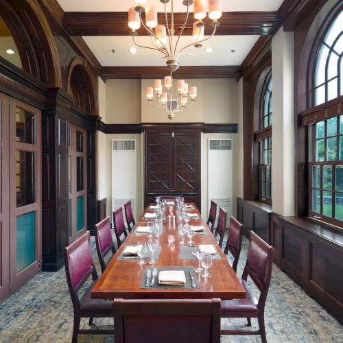 The image shows an elegant dining room with a long wooden table, set with plates and glasses, under a chandelier with arched windows.