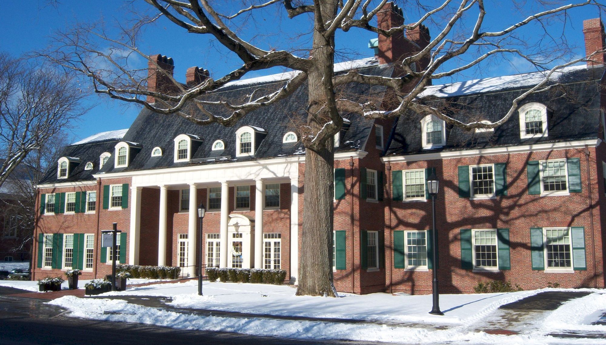 The image shows a large brick building with white columns, green shutters, and a snowy landscape under a clear blue sky.