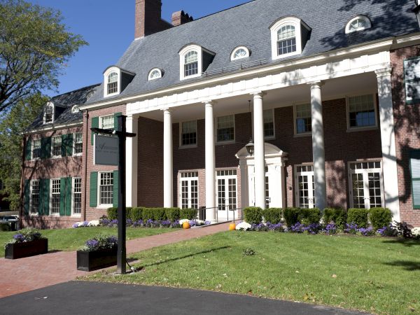 A grand brick campus building with white columns, a slate roof, green shutters, and a manicured lawn in front, under a sunny sky.